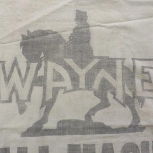 May include: A white textile featuring a faded grey illustration of a rider on horseback, with the name "WAYNE" in large letters above the horse. The text "ALL MACH" is below the horse. The fabric has a slightly creased appearance.