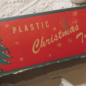 Peut inclure: Une boîte vintage pour un sapin de Noël en plastique. La boîte est rouge avec des bordures vertes et présente un sapin de Noël stylisé ainsi que le texte "Plastic Christmas Tree".