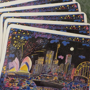 May include: A set of six colorful placemats featuring a nighttime cityscape of Sydney, Australia. The placemats have a black background with a colorful cityscape, fireworks, and stars. The text "Sydney" is written in a colorful font at the top of the placemat.