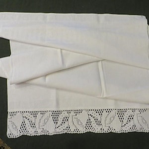 May include: A rectangular white fabric runner featuring a smooth, solid-colored center section and a wide, decorative border along the bottom edge. The border consists of intricate, hand-worked white crochet lace with a repeating leaf pattern.