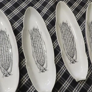 May include: Four white, oblong serving dishes with a black corn graphic. The dishes are arranged on a black and white checkered tablecloth. The corn illustration is detailed and centered on each dish.