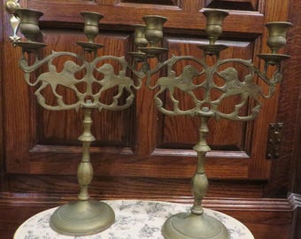 May include: Two antique brass candelabras with a decorative lion design. Each candelabra has multiple arms for candles and a round base. The candelabras are displayed on a patterned surface against a dark wood background.