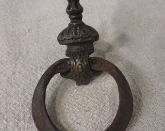 May include: A vintage, ornate, cast iron ring pull with a decorative finial. The ring is rusty and has a dark brown patina.