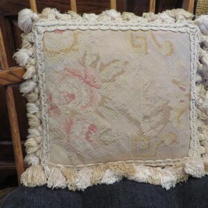 May include: A square decorative pillow featuring a needlepoint floral pattern in muted shades of pink, yellow, and cream. The pillow is bordered by a thick, braided cream-colored trim and finished with a dense fringe of matching cream tassels.