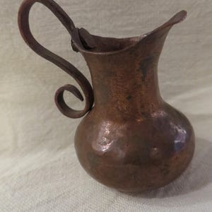May include: A small, copper pitcher with a hammered finish and a curled handle. The pitcher has a rounded body and a slightly flared lip.