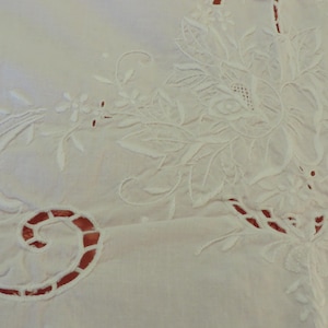 May include: Close-up of a white embroidered textile with floral and scroll designs. The embroidery is detailed with cut-out sections revealing a red fabric underneath. The overall design is delicate and intricate, suggesting a vintage or handmade item.