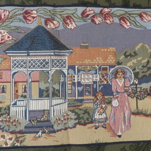 May include: Tapestry depicting a quaint town scene with a gazebo, shops, and figures. The artwork features a color palette of blues, pinks, and greens, with floral accents and a border. The words "Quilt Shop" and "General Store" are visible.