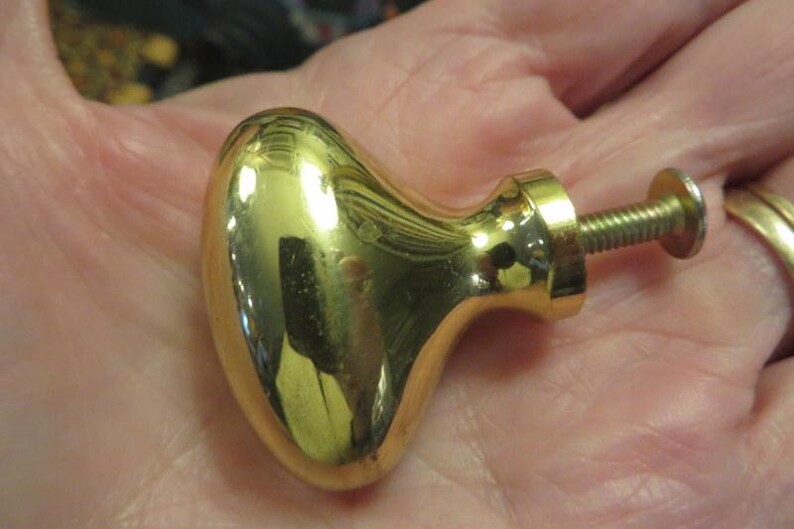 May include: A polished gold-tone metal cabinet knob with an oval, rounded head and a threaded metal screw post extending from the back. The surface of the knob is highly reflective, showing light reflections and a smooth, curved finish.