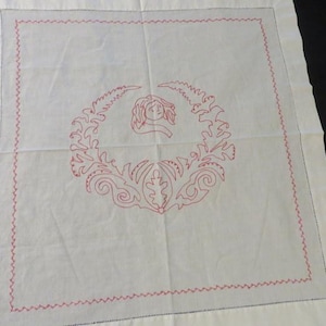 May include: White square textile with a red embroidered design. The design features a central face within a wreath of leaves and floral elements. A red zig-zag stitch borders the square.
