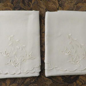 May include: Two folded white linen pillowcases with embroidered floral designs. Each pillowcase features a scalloped edge and a delicate embroidered floral pattern in the corner. The pillowcases are displayed on a patterned surface.