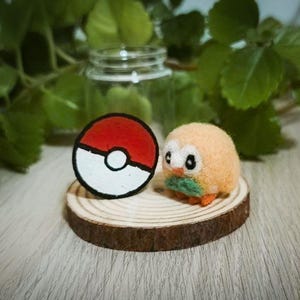 Op de afbeelding: Een naaldgevilte beige uilenfiguur met een groene vlinderdas en oranje voeten, geplaatst naast een cirkelvormig rood en wit Poké Ball-embleem op een ronde houten schijf, met een doorzichtige glazen pot op de achtergrond.