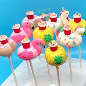 May include: A collection of colorful cake pops on sticks, featuring flamingo, pineapple, and unicorn designs. Each cake pop is topped with a miniature red cup filled with frosting and sprinkles. The background is a solid light blue.