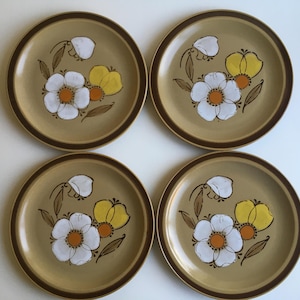May include: Set of four brown stoneware dinner plates with a floral design. The plates feature white flowers with yellow centers and brown stems on a tan background.