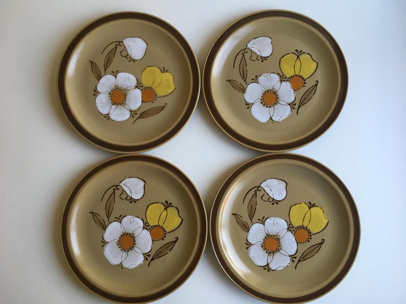 May include: Set of four brown stoneware dinner plates with a floral design. The plates feature white flowers with yellow centers and brown stems on a tan background.
