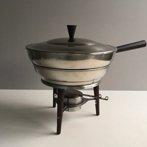 May include: A silver chafing dish with a wooden handle and a stand with three wooden legs. The chafing dish has a lid with a wooden knob.