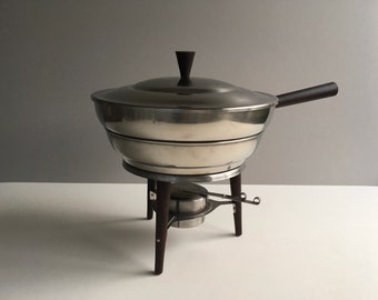 Op de afbeelding: Een zilveren chafing dish met een houten handvat en een standaard met drie houten poten. De chafing dish heeft een deksel met een houten knop.
