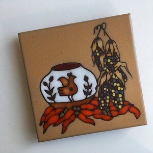 May include: A brown ceramic tile with a white pot with a brown bird design, red chili peppers, and a brown and yellow corn stalk.