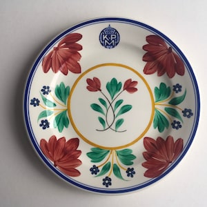 May include: A white ceramic plate with a blue rim and floral design. The center of the plate features a yellow circle with a green floral design. The rim of the plate has a red floral design with blue accents. The plate has the initials "K.P.M." in blue.