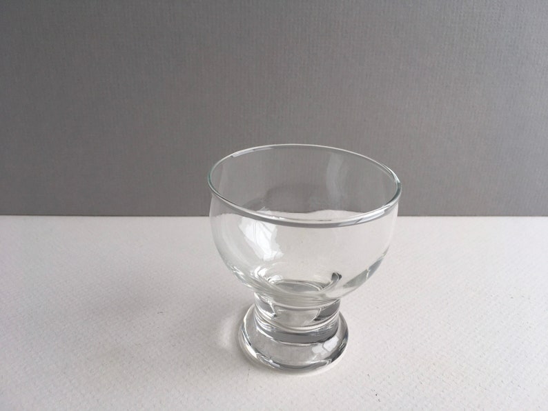 May include: A clear glass bowl with a wide base and a rounded top. The bowl is empty and sits on a white surface.