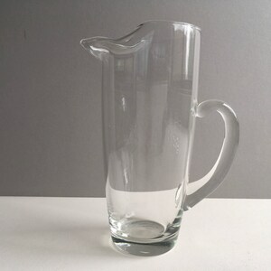 May include: A clear glass pitcher with a handle. The pitcher is tall and cylindrical with a slightly flared lip.