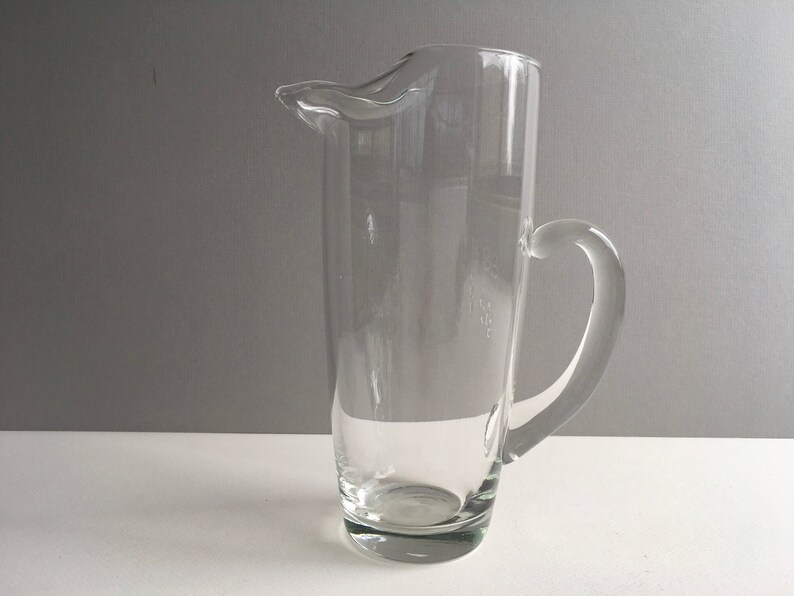 May include: A clear glass pitcher with a handle. The pitcher is tall and cylindrical with a slightly flared lip.