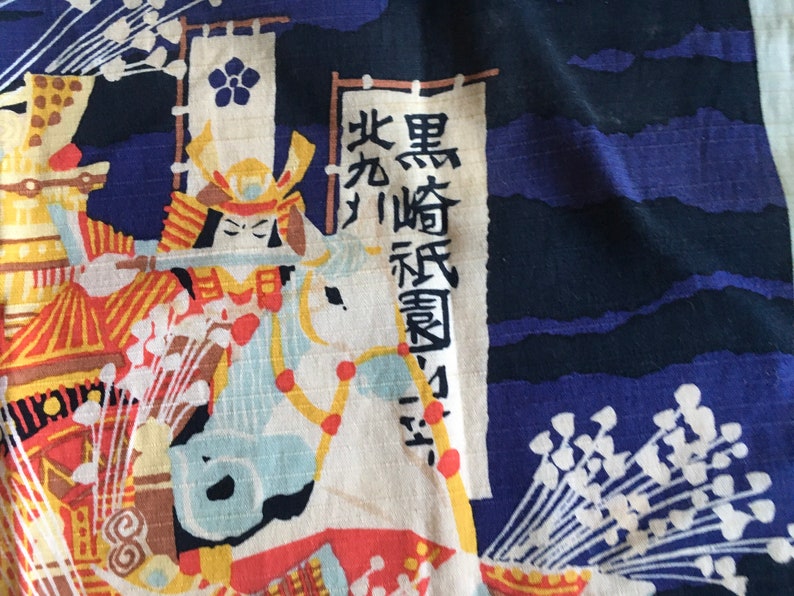 May include: A blue and white fabric featuring a Japanese warrior riding a horse. The warrior is wearing a helmet and armour. The fabric has a white flower design and the text "黒崎 辰 勇" in Japanese.