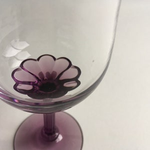 Op de afbeelding: Een helder glazen wijnglas met een paars bloemenontwerp op de bodem van de kom. De steel en voet van het glas zijn ook paars.