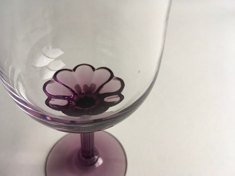 Op de afbeelding: Een helder glazen wijnglas met een paars bloemenontwerp op de bodem van de kom. De steel en voet van het glas zijn ook paars.
