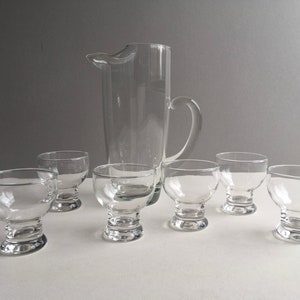 May include: A set of six clear glass drinking glasses, including a pitcher, with a wide base and a rounded bowl. The glasses are arranged on a white surface.