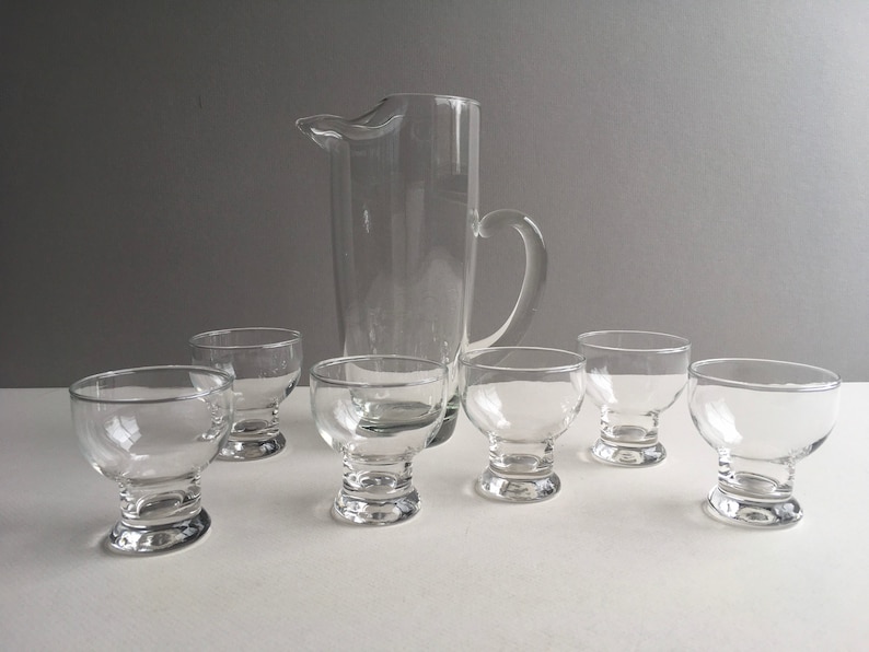May include: A set of six clear glass drinking glasses, including a pitcher, with a wide base and a rounded bowl. The glasses are arranged on a white surface.