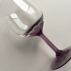 Op de afbeelding: Een helder glazen wijnglas met een paarse steel en voet. Het glas heeft een uniek ontwerp met een reeks kleine, verhoogde bulten rond de rand.