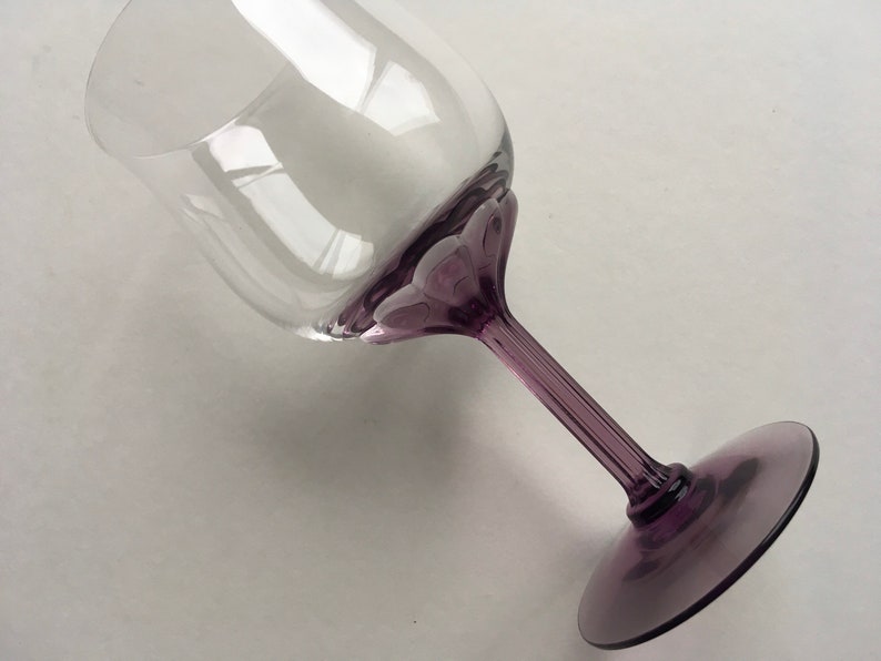 Op de afbeelding: Een helder glazen wijnglas met een paarse steel en voet. Het glas heeft een uniek ontwerp met een reeks kleine, verhoogde bulten rond de rand.