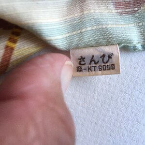 May include: A white fabric with a colourful pattern and a white tag with the text "さんび" and "番-KT 6059" in black.