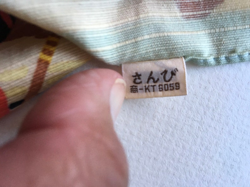May include: A white fabric with a colourful pattern and a white tag with the text "さんび" and "番-KT 6059" in black.