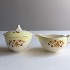 May include: A vintage cream-coloured sugar bowl and creamer set with a yellow and white ombre glaze. The set features a delicate floral design with red berries.