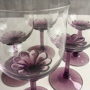 May include: Set of five clear glass wine glasses with purple flower-shaped bases. The glasses are arranged in a semi-circle.