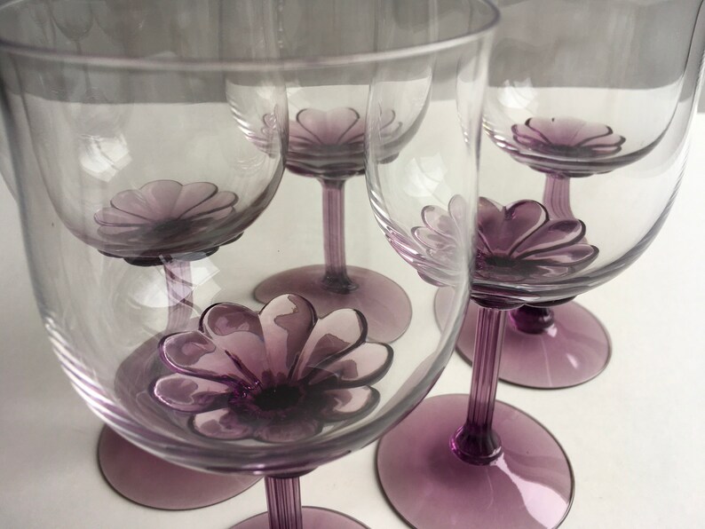 May include: Set of five clear glass wine glasses with purple flower-shaped bases. The glasses are arranged in a semi-circle.