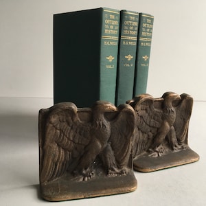 May include: Antique bronze-colored bookends featuring a detailed eagle design. Three green hardcover books with gold lettering and the title "The Outline of History" stand behind the bookends. A classic and decorative set.