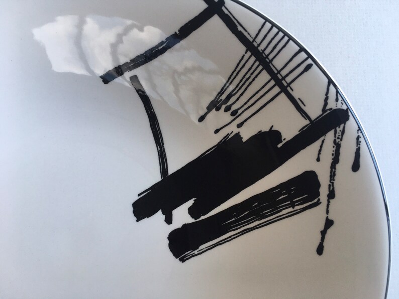 May include: A white ceramic bowl with a black and white abstract design. The design features bold brushstrokes and splatters of black paint.