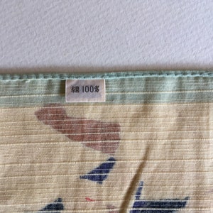 May include: A close-up of a light green and beige fabric with a brown and blue pattern. A white tag with black text reads "綿 100%".