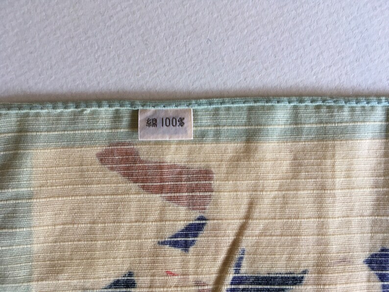 May include: A close-up of a light green and beige fabric with a brown and blue pattern. A white tag with black text reads "綿 100%".