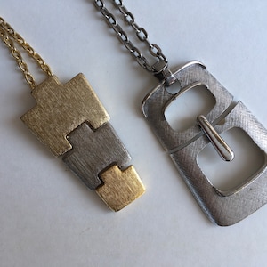 May include: Two silver necklaces with different pendants. One pendant is a geometric shape made of silver and gold metal. The other pendant is a silver buckle shape.