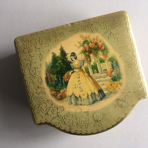 May include: A vintage gold-coloured metal box with a decorative floral border. The lid depicts a painted scene of a woman in a yellow dress in a garden. The box has a unique, curved shape and is likely a collectible item.