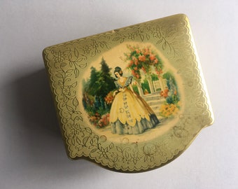 Op de afbeelding: Een vintage goudkleurige metalen doos met een decoratieve bloemenrand. Het deksel toont een geschilderde scène van een vrouw in een gele jurk in een tuin. De doos heeft een unieke, gebogen vorm en is waarschijnlijk een verzamelobject.