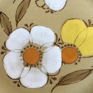 May include: A close-up of a brown ceramic plate with a floral design. The plate features white flowers with orange centers and yellow flowers with brown stems.