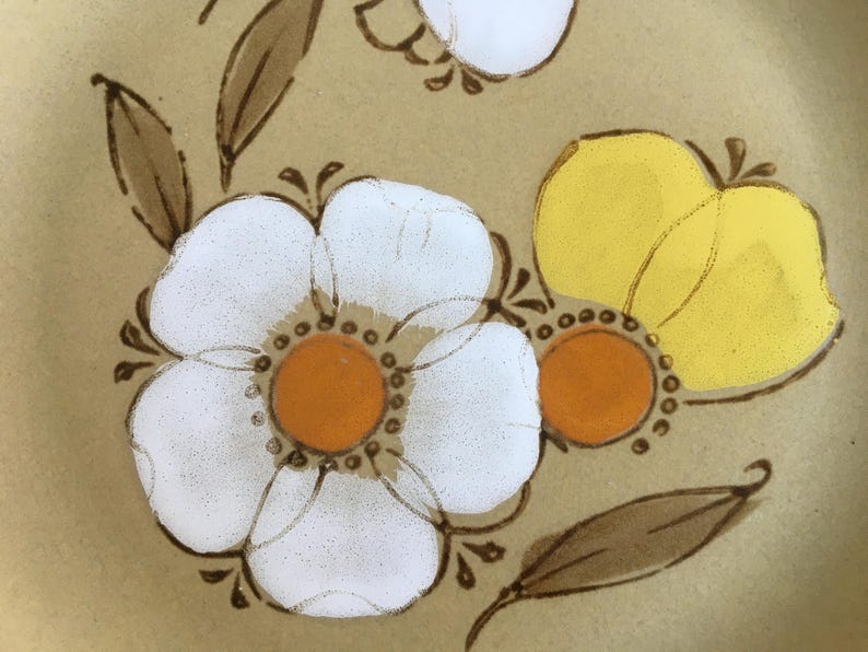 May include: A close-up of a brown ceramic plate with a floral design. The plate features white flowers with orange centers and yellow flowers with brown stems.