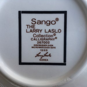 May include: A white ceramic plate with a black and white border. The plate has a brown inscription that reads "Sango&reg; THE LARRY LASLO Collection CALLIGRAPHY&reg; 267002 DISHWASHER OVEN MICROWAVE OVEN SAFE 1986 Larry Laslo KOREA".