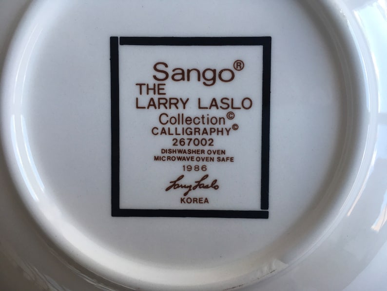 May include: A white ceramic plate with a black and white border. The plate has a brown inscription that reads "Sango&reg; THE LARRY LASLO Collection CALLIGRAPHY&reg; 267002 DISHWASHER OVEN MICROWAVE OVEN SAFE 1986 Larry Laslo KOREA".