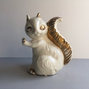 May include: A white ceramic squirrel figurine with a gold-painted tail. The squirrel has large, black eyes and a whimsical expression.