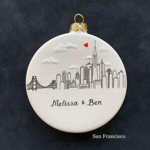 May include: White ceramic Christmas ornament with a black and white illustration of the San Francisco skyline, including the Golden Gate Bridge. The ornament also features the text "Melissa & Ben" and "San Francisco".